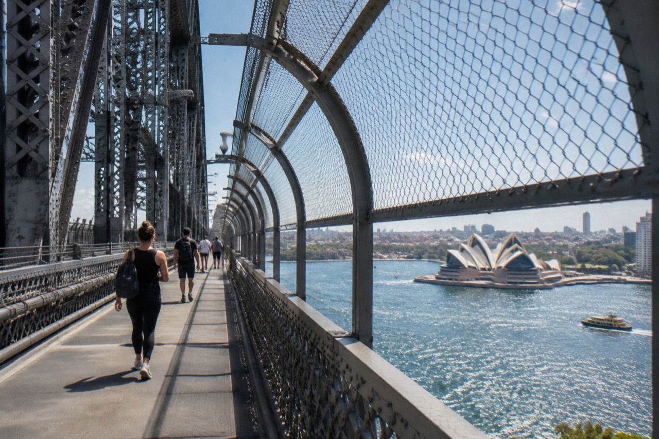 Pedestrians walking the Sydney Harbour Bridge footpath high above the harbour, with the Opera House visible below to the east