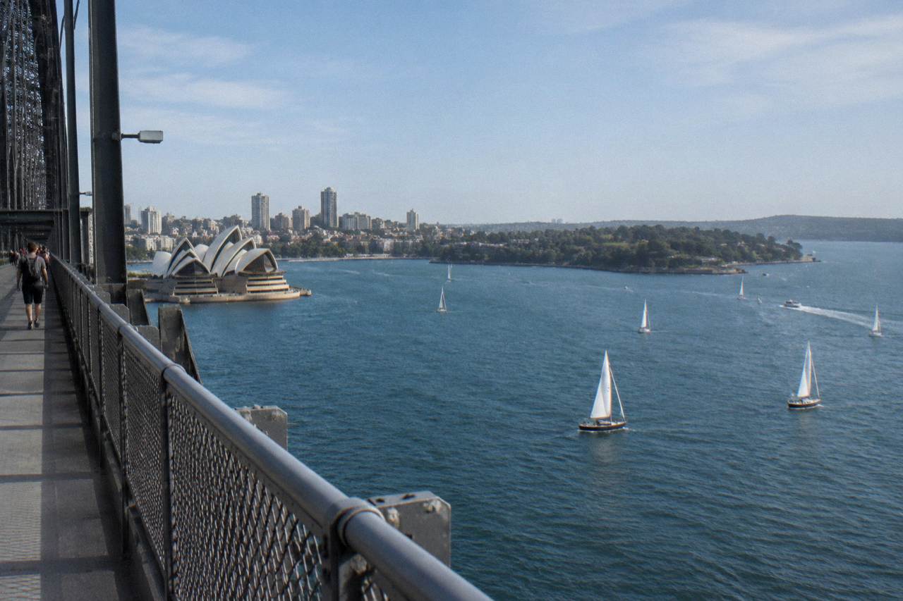 The Sydney Harbour Bridge seen from the Circular Quay foreshore, its steel arch spanning the harbour with the Opera House roofline in the distance.